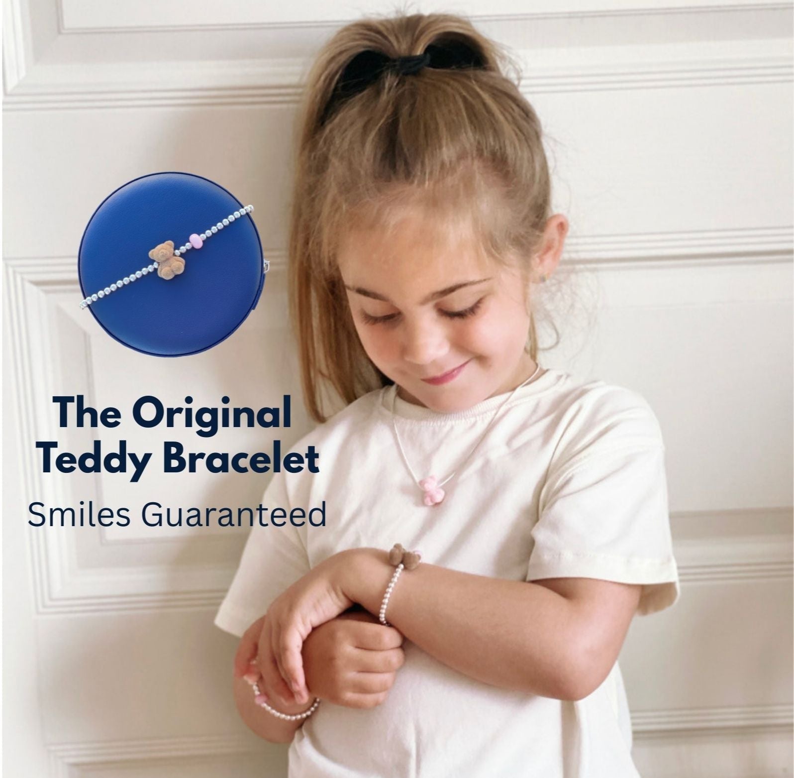 Young girl wearing a teddy bracelet with a blue box labeled 'The Original Teddy Bracelet' on a white wall.